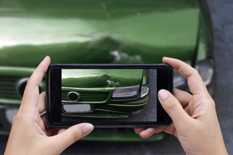 What to do if Debris hits your car - Hands holding a cell phone taking a photo of a dent in the bumper of a green car.