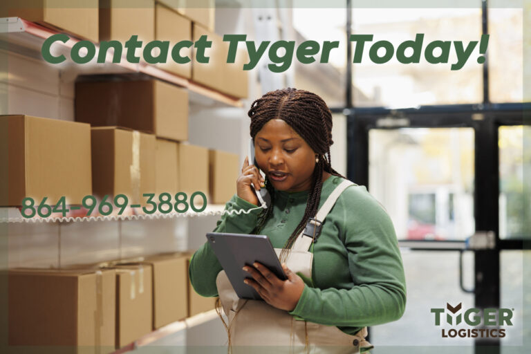 Contact Tyger Logistics Company in Woodruff, SC for reliable logistics solutions in the USA and Canada. 13101 US-221, Woodruff, SC 29388. Woman in green shirt and overalls is on a phone and tablet in a warehouse full of boxes with glass doors.